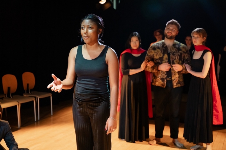 Actor stands with right hand stretched out in mid-speech while three other actors stand behind her, focused on her performance.